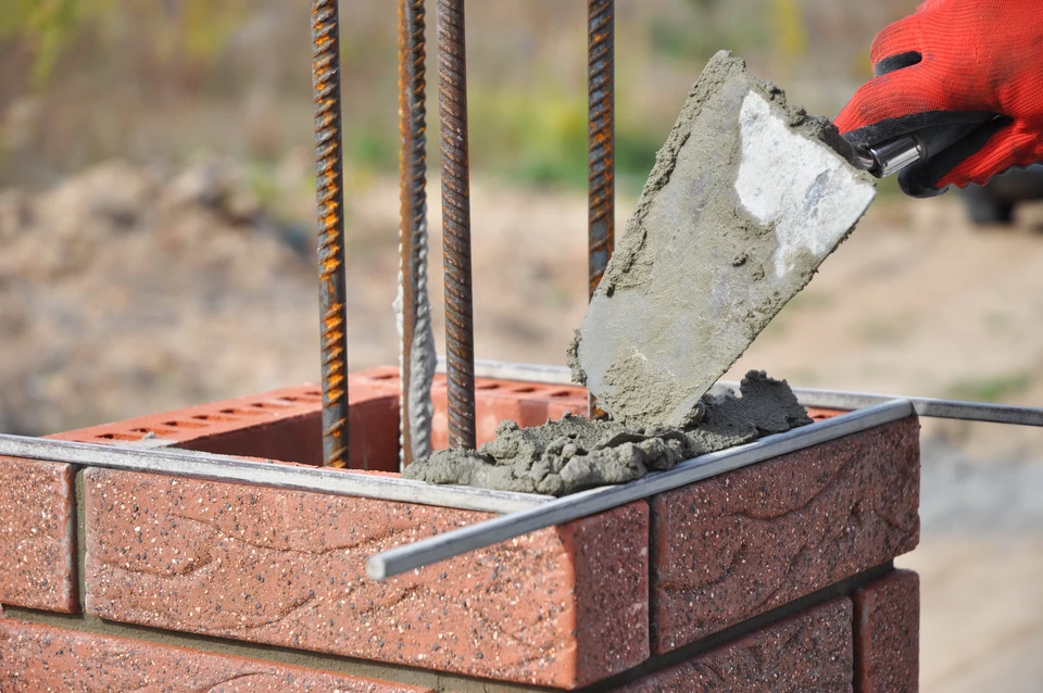 Chantier de construction en extérieur, ouvrier en gants rouges coulant du béton frais avec une truelle dans un moule délimité par des briques rouges perforées