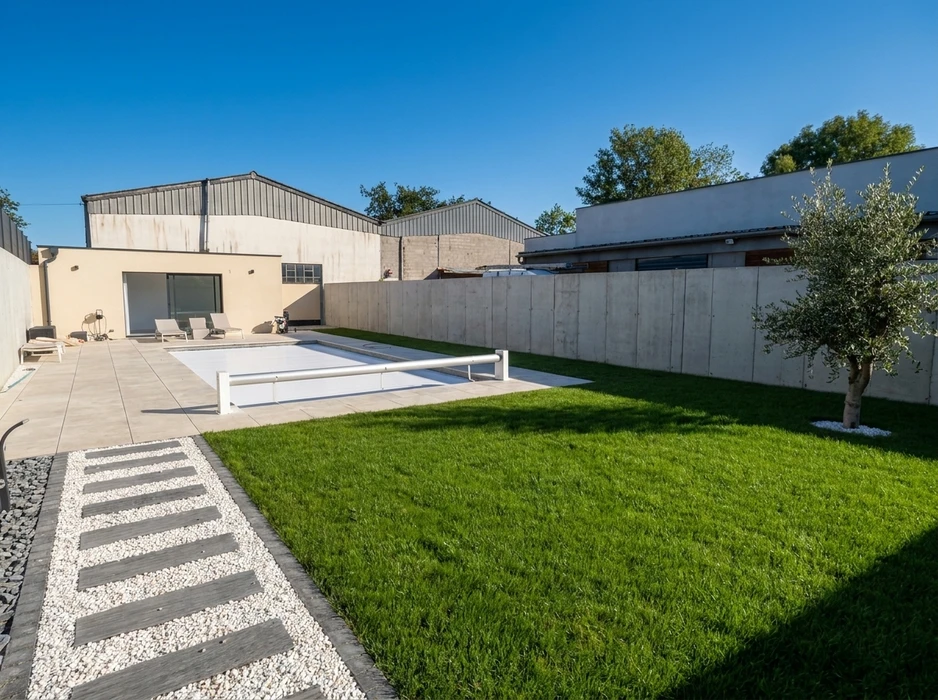 Jardin privatif moderne avec pelouse soignée, piscine couverte d'un volet roulant automatique blanc, terrasse en dalles grès cérame grand format